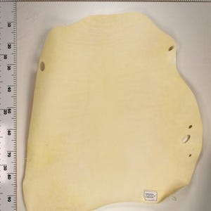 Puede incluir: Un trozo de cuero amarillo pálido con bordes irregulares y varios agujeros pequeños. El cuero está colocado sobre una superficie blanca, con una regla a lo largo del borde izquierdo que muestra medidas en centímetros. Una pequeña etiqueta es visible.