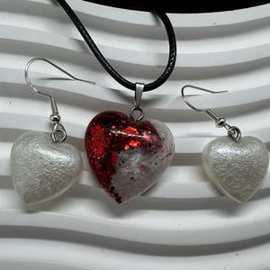 May include: A heart-shaped necklace and earrings set. The necklace and earrings are made of silver and have a white heart pendant with red glitter. The necklace has a black cord.