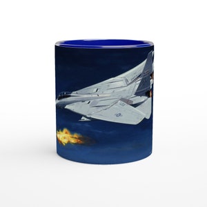May include: A white and gray fighter jet with blue and orange flames coming out of the back of the jet. The jet is flying in a dark blue sky. The image is on a blue and white coffee mug.
