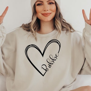 May include: Beige sweatshirt with a black heart design and the word "Bubbe" in script. The sweatshirt is paired with a matching beige beanie. The model is giving a peace sign.