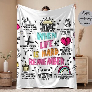 May include: White blanket with colourful text and illustrations. The blanket features phrases like "When Life is Hard Remember..." and Bible verses. Designs include a butterfly, rainbow, and hearts. The blanket is held up by a person.