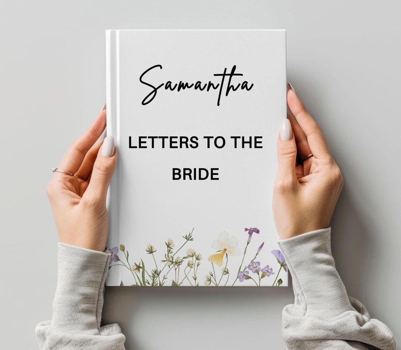 May include: A white journal titled "Samantha LETTERS TO THE BRIDE" in black script. The journal features a floral design along the bottom edge with purple, yellow, and white flowers. The journal is held by two hands.