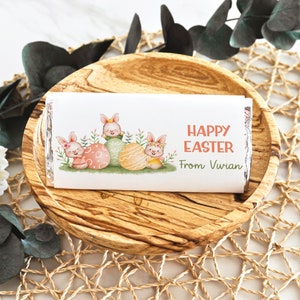 May include: A white chocolate bar wrapper with a design of three bunnies holding Easter eggs. The wrapper has the text "Happy Easter" and "From Vivian" printed on it.