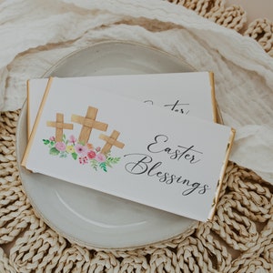 May include: A white chocolate bar with a gold foil wrapper. The bar is decorated with a watercolor illustration of three wooden crosses with pink and yellow flowers. The text "Easter Blessings" is written in black cursive script.