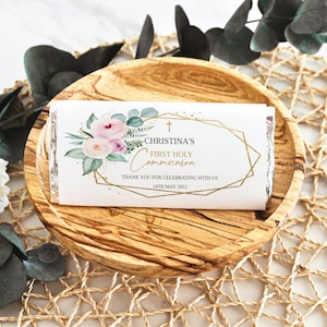 May include: A white chocolate bar wrapper with floral and geometric gold accents. The wrapper reads "Christina's First Holy Communion" with a cross and the date "14th May 2023". The bar rests on a wooden plate.