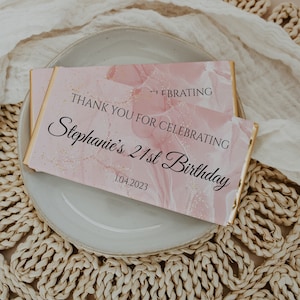 May include: Two pink and gold birthday chocolate bars with the text "THANK YOU FOR CELEBRATING Stephanie's 21st Birthday 1.04.2023". The bars are on a white plate, with a woven placemat in the background.