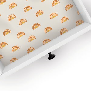 May include: White drawer with a black knob. The inside of the drawer is lined with a cream-colored wallpaper featuring a repeating pattern of orange sunbursts.