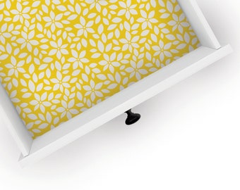 May include: An open white drawer with a black knob displays a yellow floral pattern lining its interior. The pattern features white, stylized flower shapes. The drawer is set against a white background.
