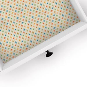 May include: A white drawer with a black knob. The inside of the drawer is lined with a patterned paper featuring a repeating design of colorful stars on a cream background.
