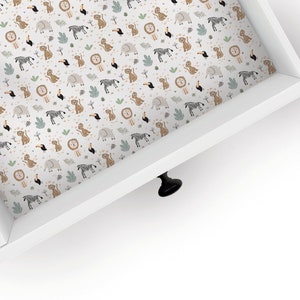 May include: A white drawer with a black knob. The inside of the drawer is lined with a white fabric with a repeating pattern of lions, zebras, elephants, toucans, and leaves.