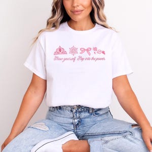 May include: White t-shirt featuring a pink graphic of a crown, snowflake, bow, and flower. The text "Show yourself. Step into the power" is printed below the graphics. The shirt is made of a soft material.