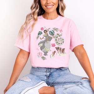 May include: Pink t-shirt featuring a colorful illustration of characters from a classic animated film. The design includes a boy riding a bear, a snake, and other jungle elements. The shirt is a soft, comfortable material.