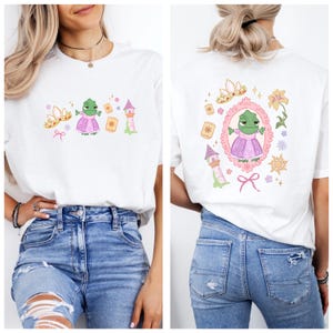 May include: White t-shirt with a cartoon frog in a pink dress, crown, tower, and lanterns. The back design mirrors the front, with the frog in a pink frame, surrounded by flowers and a sun.