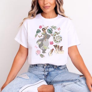 May include: White t-shirt featuring a colorful illustration of a cartoon scene with a bear, a girl, a snake, and other characters. The design includes pink flowers and green leaves. The shirt is made of a soft material.