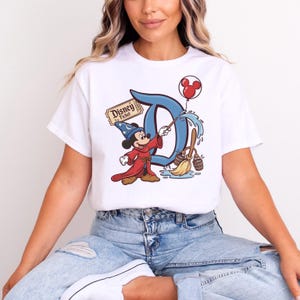 May include: White t-shirt with a colourful graphic of Mickey Mouse as a sorcerer, with the word "Disney" and a ticket. Mickey is holding a wand, with a blue "D" behind him. A red Mickey Mouse balloon is in the upper right.