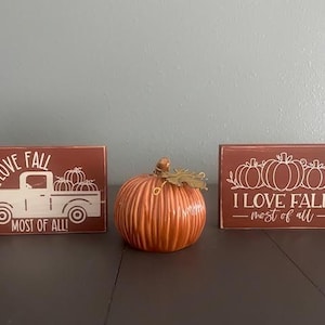May include: Three wooden signs with the text "I love fall - most of all" and a decorative ceramic pumpkin. The signs are painted in a reddish-brown color and have white lettering. The pumpkin is orange and has a brown stem.