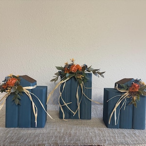 May include: Three decorative stacks of books painted in a matte dark blue finish. Each stack is topped with an arrangement of artificial orange flowers, green leaves, and dried blue accents, tied together with thin strands of natural raffia ribbon.