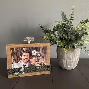 May include: A wooden picture frame with a photo of a couple, a butterfly embellishment, and a decorative knob on top. The frame is sitting on a dark brown surface.