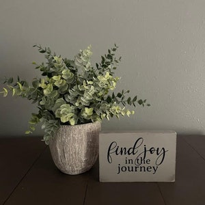 May include: A wooden sign with the text "find joy in the journey" in black lettering on a light gray background. A potted plant with green leaves is in the background.
