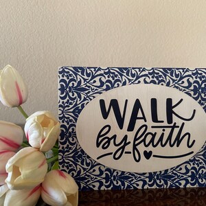 May include: A wooden sign with the words "WALK by faith" in black script on an oval, set against a blue and white floral pattern. A bouquet of white tulips with pink accents is in the foreground.