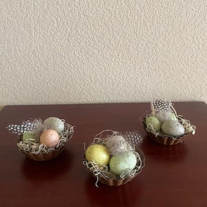 May include: Three small brown metal nests filled with faux bird nests, feathers, and speckled pastel green, yellow, and pink Easter eggs.