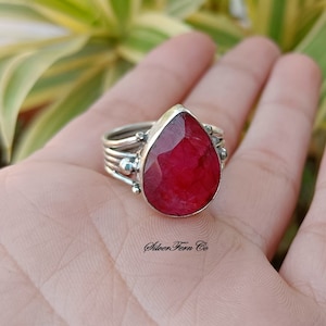 Può includere: Un anello d'argento con una grande pietra preziosa di rubino a forma di goccia. L'anello ha una fascia delicata a più livelli con piccole perle d'argento.