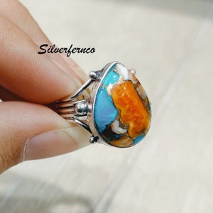 May include: A silver ring featuring a teardrop-shaped turquoise and orange gemstone. The stone displays a distinctive pattern of turquoise, orange, and white.
