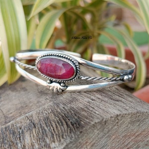 Peut inclure: Un bracelet en argent avec une pierre rouge sertie dans un ovale en argent. Le bracelet a un design de fil torsadé et un petit détail décoratif sur le côté.