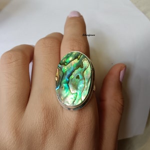 May include: A silver ring with a large oval abalone shell inlay. The shell has a green, blue, and iridescent color pattern.