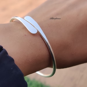 Può includere: Un braccialetto rigido in argento con un design unico, caratterizzato da una fascia curva e un pezzo piatto e angolato che si avvolge attorno alla fascia.