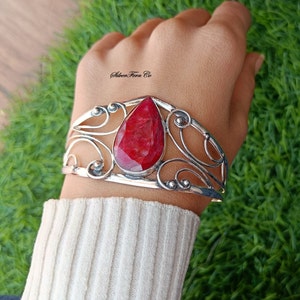May include: A silver cuff bracelet featuring a large, red ruby gemstone set in the centre. The bracelet has a delicate, ornate design with swirls and curves.