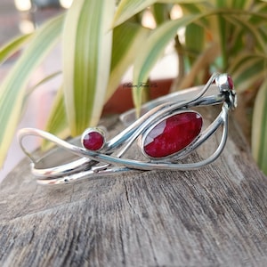 Puede incluir: Un brazalete de plata con tres acentos de piedras preciosas rojas. El brazalete tiene un diseño delicado y orgánico con un patrón de vid.