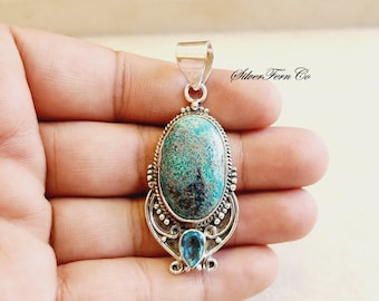 May include: A silver pendant with a large oval turquoise stone set in a silver filigree design. A smaller blue topaz stone is set below the turquoise stone. The pendant is on a hand, and the text "Silver Fern Co." is visible in the background.