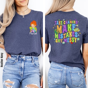 May include: A heather navy blue t-shirt with a cartoon teacher and the text "Miss Karl" on the front. The back of the shirt has the text "Take Chances Make Mistakes Get Messy" with colorful illustrations of school supplies.
