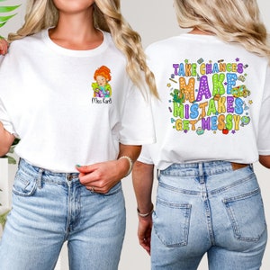 May include: White t-shirt with a cartoon teacher illustration and the text "Miss Kari" on the front. The back of the shirt has the text "Take Chances Make Mistakes Get Messy" with colorful school supplies and stars.