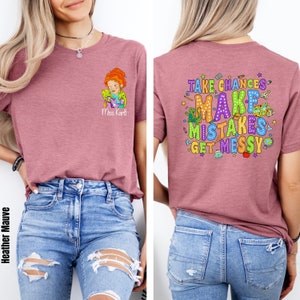 May include: A heather mauve t-shirt with a cartoon image of a teacher holding a lizard on the front and the text "Take Chances, Make Mistakes, Get Messy" on the back. The shirt is being modeled by a person wearing blue jeans.