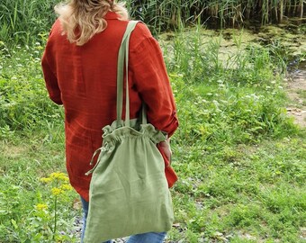 May include: A person wearing a rust-colored linen shirt and blue jeans, carrying a light green drawstring tote bag. The bag has long straps and a gathered top. The background features lush green foliage and a body of water.
