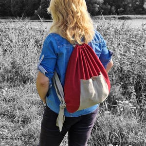 May include: A woman wearing a blue denim shirt and black trousers is carrying a red and beige drawstring backpack. The backpack has a small pocket on the front.