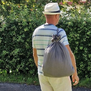 Könnte beinhalten: Eine Person mit einem weißen Hut und einem gestreiften Hemd trägt eine graue Kordeltasche über der Schulter. Die Person geht auf einem Weg in der Nähe einer grünen Hecke.
