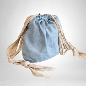 Puede incluir: Una bolsa de cordón de color azul claro con correas de color crema. La bolsa está hecha de un tejido de lino suave.