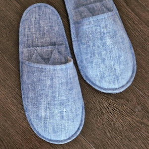 May include: Two blue linen slippers with a quilted design on a wooden surface. The slippers have a rounded toe and a soft, comfortable appearance. The color is a light, faded blue.