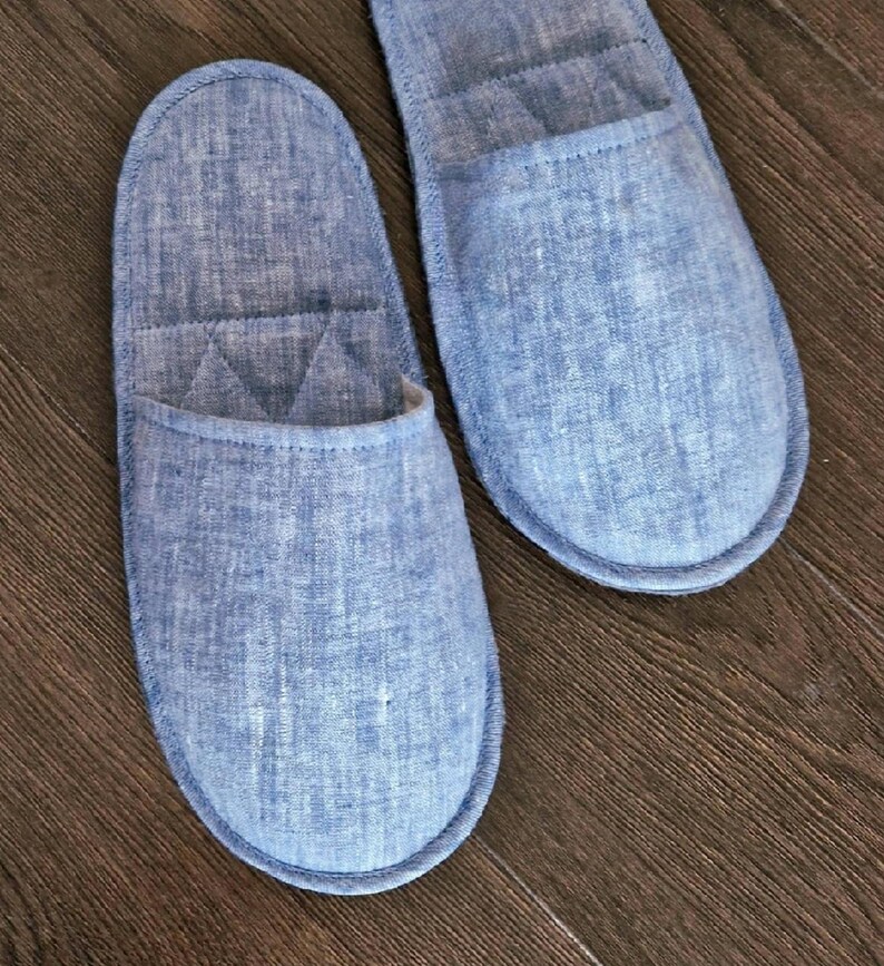 May include: Two blue linen slippers with a quilted design on a wooden surface. The slippers have a rounded toe and a soft, comfortable appearance. The color is a light, faded blue.
