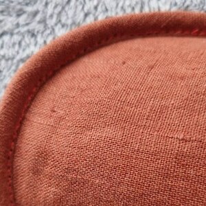 May include: Close-up of a rust-colored, rounded fabric item with a stitched edge. The fabric has a textured appearance, and the stitching is a slightly darker shade of the same color. The background is a soft, gray, fuzzy material.