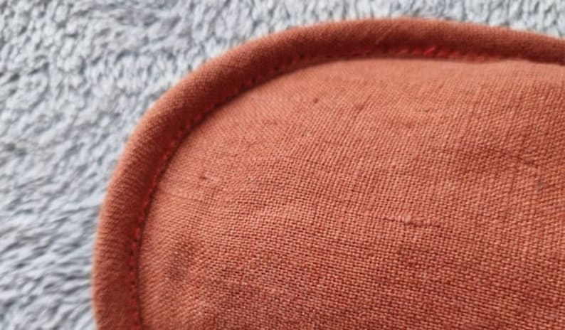 May include: Close-up of a rust-colored, rounded fabric item with a stitched edge. The fabric has a textured appearance, and the stitching is a slightly darker shade of the same color. The background is a soft, gray, fuzzy material.