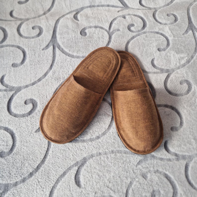 May include: A pair of brown, open-toe slippers. The slippers are made of a soft, textured material and have a simple, classic design. They are resting on a light gray patterned surface.