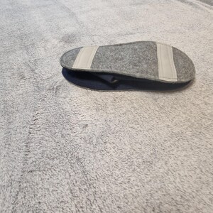 May include: A gray felt slipper insole with two white fabric strips is placed on top of a dark blue slipper. The insole is oval-shaped and appears to be made of a soft, textured material. The slipper is resting on a gray, fuzzy surface.