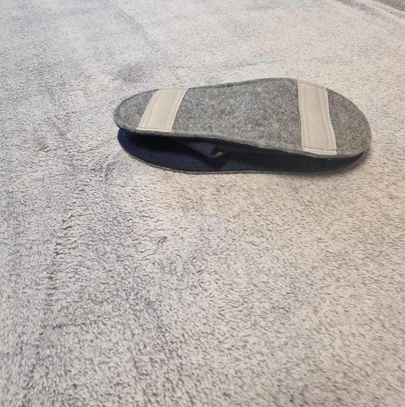 May include: A gray felt slipper insole with two white fabric strips is placed on top of a dark blue slipper. The insole is oval-shaped and appears to be made of a soft, textured material. The slipper is resting on a gray, fuzzy surface.