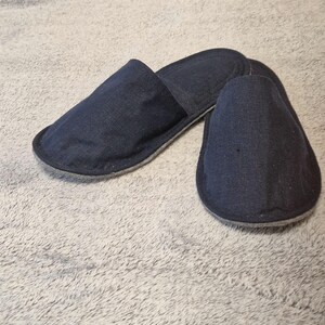 May include: A pair of dark blue denim slippers. The slippers have a simple design with a rounded toe and a slip-on style. The soles are a light gray color, and the slippers are made of a soft, comfortable material.