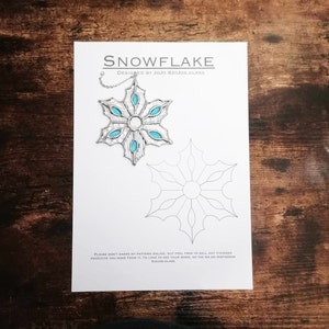 Puede incluir: Un patrón imprimible para un adorno de copo de nieve de vitral. El patrón está delineado en negro sobre un fondo blanco. El adorno terminado se muestra en la esquina superior izquierda, con un marco plateado y piezas de vidrio turquesa.
