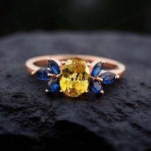 Puede incluir: Un anillo de oro rosa con una gran piedra preciosa amarilla ovalada rodeada de piedras preciosas azules más pequeñas.